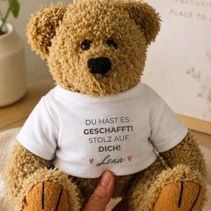 Könnte beinhalten: Ein brauner Plüsch-Teddybär, der ein weißes T-Shirt trägt. Auf dem T-Shirt steht der Text "DU HAST ES GESCHAFFT! STOLZ AUF DICH!", gefolgt von dem Namen "Lena", der von zwei kleinen rosa Herzen flankiert wird. Der Bär hat dunkle Augen und eine schwarze Nase.