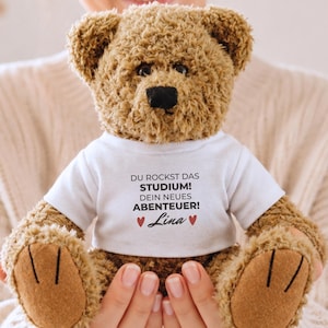 Könnte beinhalten: Ein brauner Plüsch-Teddybär in einem weißen T-Shirt. Auf dem T-Shirt steht der Text "DU ROCKST DAS STUDIUM! DEIN NEUES ABENTEUER!" mit dem Namen "Lina" in Schreibschrift zwischen zwei kleinen roten Herzen auf der Vorderseite gedruckt.