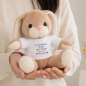 Può includere: Un coniglio di peluche marrone chiaro con orecchie lunghe e naso rosa, che indossa una maglietta bianca stampata con il testo tedesco 'Ein Stück Heimat für dein neues Zuhause', il nome 'Emma 2026' e due icone a forma di cuore rosso.