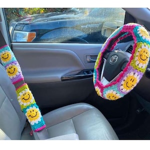 May include: Crocheted car accessories set featuring a steering wheel cover and seatbelt covers. The set is decorated with a colorful daisy and smiley face pattern. The color palette includes yellow, pink, blue, green, and purple.