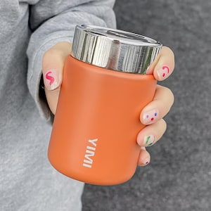 May include: An orange insulated tumbler with a silver lid is held in a person's hand. The tumbler has the word "YIMI" printed vertically in white. The person's nails are painted with colorful designs.