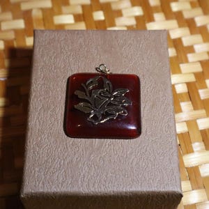May include: A square, red pendant with a gold floral design, resting on a textured, tan box. The pendant has a gold bail. The background is a woven, light brown surface.