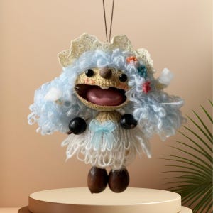 May include: A handmade crochet doll with a whimsical design. The doll features blue yarn hair, a wide open mouth, and a light blue bow. The doll is adorned with small flowers and black bead accents. The doll is displayed on a round, light brown platform.