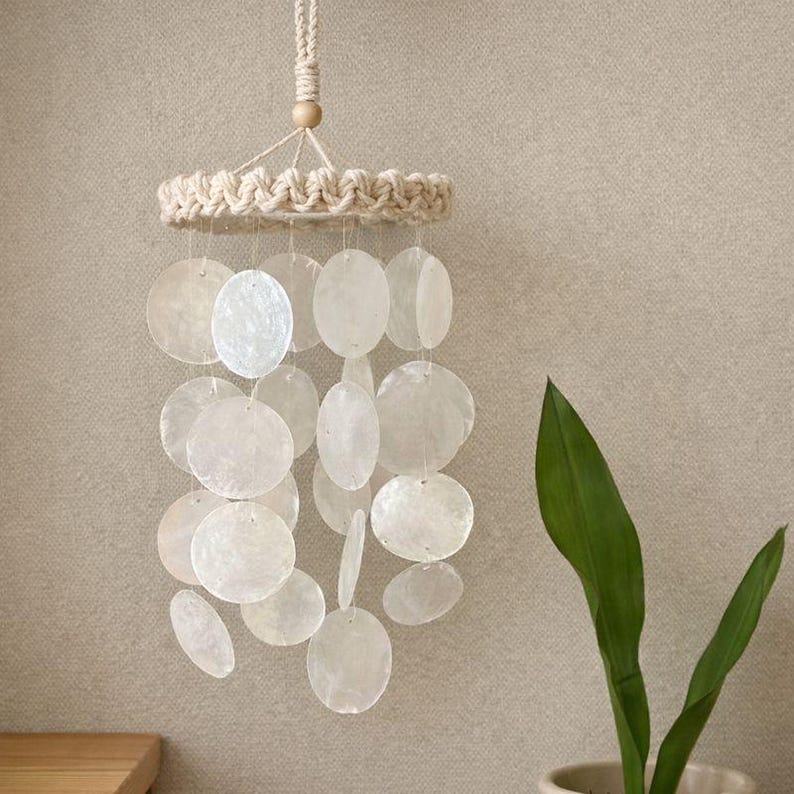 May include: A hanging wind chime featuring a macrame top and numerous white, oval-shaped shell discs. The discs are suspended from the macrame ring by thin, clear strings. A wooden bead is at the top of the chime. The wind chime is approximately 12 inches long.
