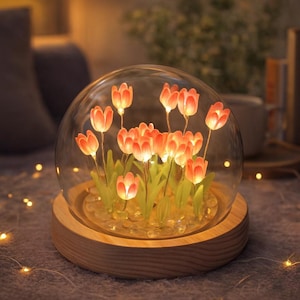 Op de afbeelding: Een decoratieve glazen koepel met verlichte oranje tulpenbloemen. De bloemen hebben groene stelen en liggen op heldere stenen. De koepel rust op een ronde, lichtgekleurde houten basis, wat een warme, omgevingsgloed creëert.