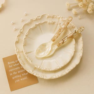 Peut inclure: Ensemble de trois assiettes crème à bord festonné et deux cuillères assorties. Les cuillères et les assiettes ont une finition nacrée. Une petite carte avec du texte est dans le coin inférieur gauche. Les assiettes sont empilées.