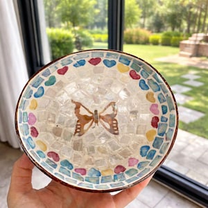 Könnte beinhalten: Dekorative Mosaikschale mit Schmetterlingsdesign. Die Schale hat einen braunen Rand und ein Mosaikmuster mit einem Schmetterling in der Mitte. Das Mosaik umfasst weiße, blaue, rote, gelbe und rosa Fliesen. Die Schale wird in einer Hand gehalten.