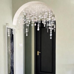 May include: A decorative archway with a cascading beaded curtain of white, iridescent, circular beads. The archway is above a black door with a gold handle. A white framed mirror hangs on the left.