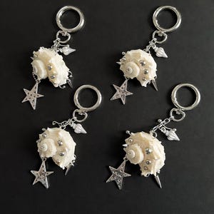 May include: Four keychains featuring cream-colored seashells adorned with silver-toned studs and charms. Each keychain has a silver star pendant and a silver ring clasp. The background is a solid black.