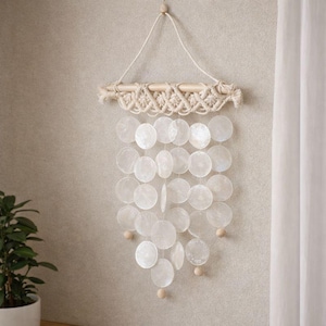 May include: A decorative wall hanging featuring a wooden dowel with macrame detailing. Below, a cascade of white, iridescent, circular shell pieces hangs, accented with small wooden beads. The piece is suspended by a natural fiber cord.