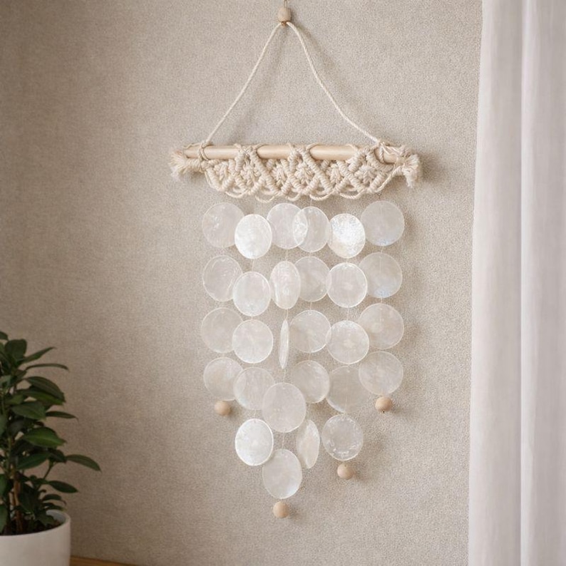 May include: A decorative wall hanging featuring a wooden dowel with macrame detailing. Below, a cascade of white, iridescent, circular shell pieces hangs, accented with small wooden beads. The piece is suspended by a natural fiber cord.