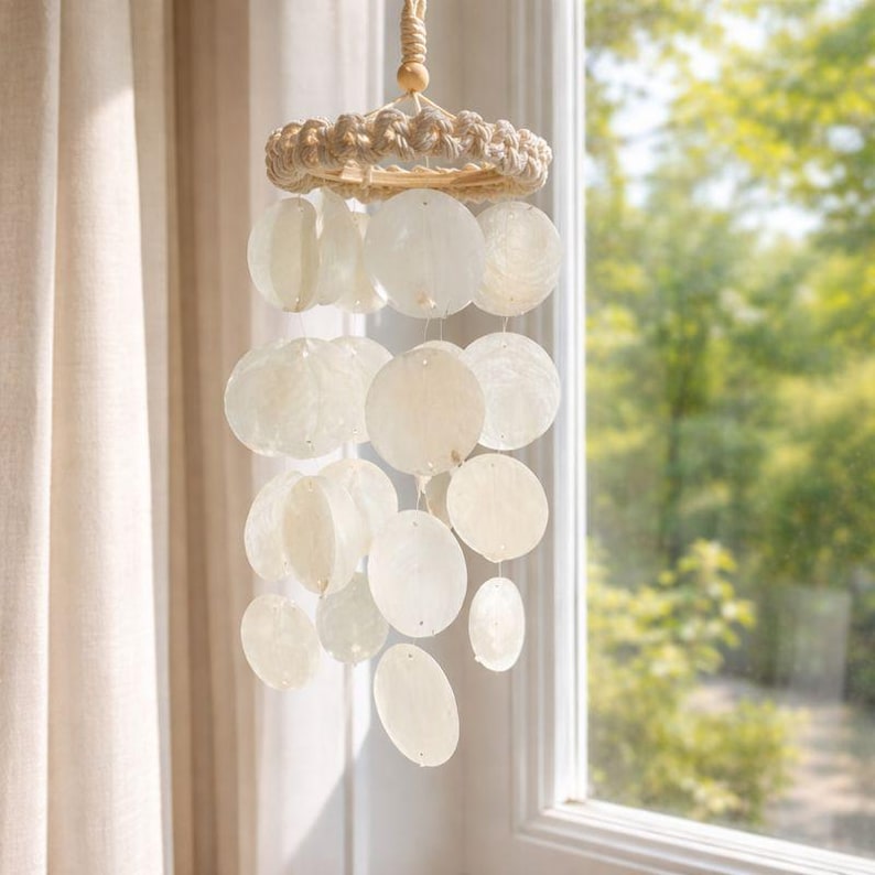May include: A decorative wind chime with a natural, coastal aesthetic. The chime features a woven ring at the top, a series of round, off-white shell discs, and a natural rope for hanging. The design is simple and elegant, suitable for indoor or outdoor use.