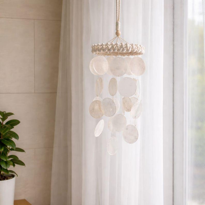 May include: A hanging wind chime with a natural, bohemian aesthetic. The chime features a woven top, a series of round, off-white shell discs, and a natural rope for hanging. The discs vary in size and hang at different lengths.