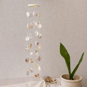 May include: A decorative wind chime made from capiz shells. The shells are in various shapes and sizes, strung together with clear thread. The shells are in natural tones of white, cream, and tan. The wind chime hangs from a wooden ring.