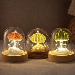 May include: Three decorative glass dome lights with wooden bases. Each dome features a sea urchin design in orange, yellow, and green, illuminated by small lights. The lights sit on a white, textured base, creating a warm glow. A unique home decor item.