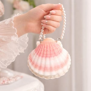 Puede incluir: Un bolso con forma de concha marina con un asa de perlas. El bolso presenta un patrón de rayas rosas y blancas, que se asemeja a una concha de vieira. El asa está hecha de perlas blancas y el bolso lo sostiene una mano con uñas cuidadas.