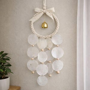 May include: A decorative wall hanging featuring a woven rope circle with a small gold bell and a cream-colored lace bow. Below, a series of translucent, round, shell-like discs are strung together with wooden beads, creating a calming, natural aesthetic.