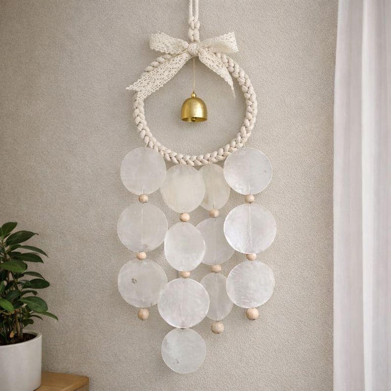 May include: A decorative wall hanging featuring a woven rope circle with a small gold bell and a cream-colored lace bow. Below, a series of translucent, round, shell-like discs are strung together with wooden beads, creating a calming, natural aesthetic.