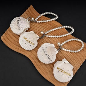 Puede incluir: Cuatro conchas de vieira blancas, cada una con un nombre o palabra en letras doradas o plateadas, incluyendo REBECCA, AMANDA, CHRISTINA y WEEDING. Tres conchas están unidas a bucles de cuentas de perlas blancas con cierres de metal plateado.