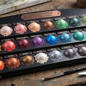 May include: A black mica powder collection with 24 vibrant colors, including pearl, gold, and copper. The powders are arranged in a tiered display with a copper-colored frame. A paintbrush and palette knives are next to the display on a wooden surface.