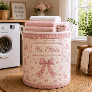 May include: A cylindrical quilted laundry hamper in a soft pink and white floral pattern with a large bow graphic. The front features an embroidered rectangular patch with the text Ms. Olivia. The hamper is filled with folded white and pink towels.