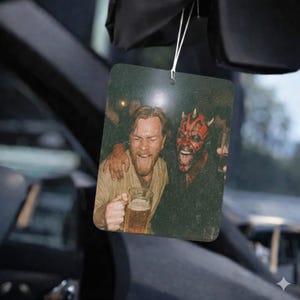 May include: Rectangular car air freshener with a vintage image of two people laughing, one holding a pint of beer. One person is wearing a red and black devil mask. The air freshener hangs from a white string.