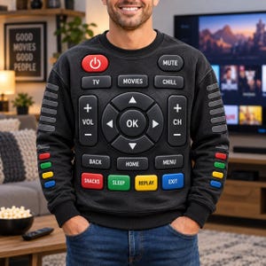 Op de afbeelding: Een zwart sweatshirt met lange mouwen en een grafische print van een afstandsbediening voor de televisie op de voorkant. Het ontwerp bevat knoppen met de labels TV, MOVIES, CHILL, VOL, CH, OK, BACK, HOME, MENU, SNACKS, SLEEP, REPLAY en EXIT.