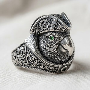 Puede incluir: Anillo de plata detallado con un loro que lleva un sombrero de pirata. El loro tiene un ojo de gema verde y un pico detallado. La banda del anillo tiene un diseño ornamental y arremolinado. El sombrero tiene detalles en forma de engranaje.