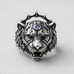 May include: A detailed silver ring shaped like a tiger's head, featuring intricate mechanical details and a purple gemstone. The ring has a crown-like design above the head and small clear stones around the eyes.