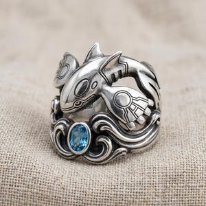 May include: A sterling silver ring featuring a sculpted creature with fins and circular markings, accented by a single oval-cut blue gemstone set within a base of swirling, wave-like metal patterns that wrap around the finger.