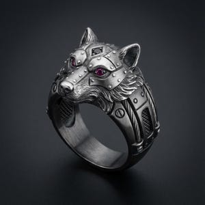 May include: A silver-toned ring featuring a wolf head design. The wolf has red gemstone eyes and is detailed with rivets and geometric patterns. The ring has a dark background.