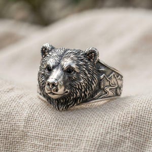 May include: A silver ring featuring a detailed, three-dimensional bear head as the centerpiece. The band is engraved with a landscape scene depicting mountains, a winding path, and pine trees. The bear has dark, inset eyes and ears.