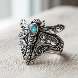 May include: A silver ring with a dragonfly motif. The ring showcases detailed craftsmanship, incorporating gears and a central oval gemstone with blue and green tones. The dragonfly's wings are finely sculpted.