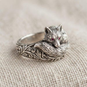 Pode incluir: Um anel de prata com uma raposa esculpida cuja cauda se enrola em torno da banda. A raposa tem duas pequenas pedras vermelhas incrustadas como olhos. A banda é decorada com intrincados relevos gravados de folhas e videiras que se estendem.