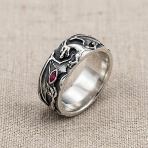 May include: A wide silver band ring featuring an embossed dragon design with oxidized detailing. The dragon is depicted with wings spread and a single marquise-cut red gemstone set into the metal surface, creating a textured and metallic finish.