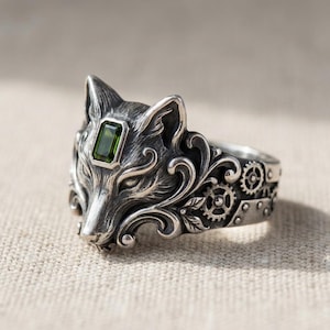 May include: A silver ring with a wolf's head design and a green rectangular gemstone. The ring showcases detailed craftsmanship, including gears and scrollwork. The wolf's head is the central element, with finely rendered fur and features.