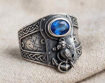 May include: A wide, silver-toned ring featuring an oval, blue gemstone. The ring has intricate nautical-themed details, including a parrot, anchor, and map design. Celtic knotwork borders the ring's edges.