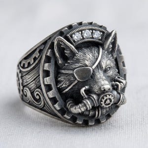 May include: A silver ring featuring a detailed wolf design. The wolf wears an eye patch and a respirator. The ring has a crown with three small gems. The ring's band has a gear and scroll design.
