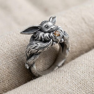 May include: A silver ring featuring a sculpted rabbit with long ears and textured fur. The rabbit holds a small, round, amber-colored gemstone at its chest. The band of the ring is formed by the rabbit's body and tail, showcasing intricate metalwork.