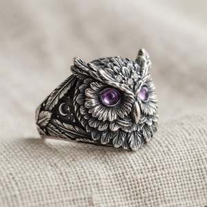Könnte beinhalten: Ein silberner Ring in Form eines Eulenkopfes mit detaillierter Federstruktur, zwei runden violetten Edelsteinaugen sowie einer kleinen Gravur von Mondsichel und Stern an der Seite. Das Metall hat ein dunkel oxidiertes Finish für Kontraste.