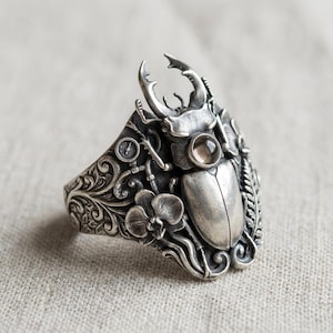 May include: A silver ring featuring a detailed beetle design. The ring has a large beetle with prominent horns, a small gemstone, and intricate floral and mechanical embellishments. The ring is set against a neutral background.