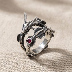 May include: A silver-toned metal ring shaped like a serpentine creature with an elongated head, segmented body, and fin-like protrusions. The design features a small, circular, deep red gemstone set into the side of the band near the creature's tail.