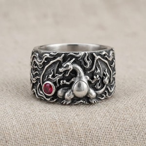 May include: A wide silver-toned ring featuring an embossed dragon-like creature surrounded by intricate flame patterns. A small, round, faceted red gemstone is set into the metal surface to the left of the central creature figure.