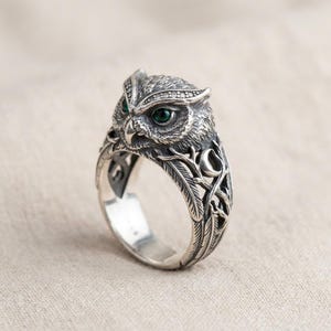 Könnte beinhalten: Ein silberner Ring mit einem detaillierten Eulenkopf, der zwei kleine grüne Steine als Augen hat. Die Ringschiene ist mit aufwendigen Gravuren von Federn, Zweigen und einer Mondsichel verziert und weist ein strukturiertes, oxidiertes Finish auf.