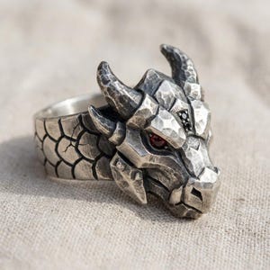 May include: A silver-toned ring featuring a sculpted dragon head with a hammered metal texture. The dragon has two prominent horns, a scale-patterned band, and a single deep red stone set as an eye, complemented by small black accent stones.
