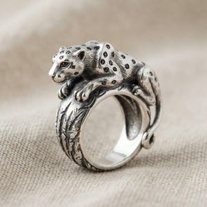 May include: A sterling silver ring featuring a sculpted leopard resting on the band. The leopard has a textured finish with small black stones embedded as spots. The band itself displays a carved, bark-like pattern around the entire circumference.