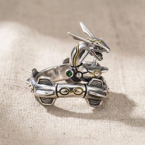 May include: A silver-toned ring shaped like a serpentine creature with an elongated head, open mouth, and pointed crest. The band features segmented armor-like plating with recessed yellow circular accents and a small, round, embedded green gemstone.