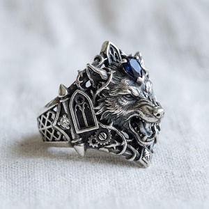 Peut inclure: Bague en argent détaillée avec une tête de loup rugissante et une pierre bleue. La bague présente des détails gothiques, une petite fenêtre, des engrenages et des épines. Un bijou unique et complexe.