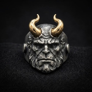 May include: A silver-toned ring featuring a detailed, textured face of a horned creature with a stern expression. The ring has two prominent, smooth, gold-toned curved horns extending from the top of the head, contrasting with the dark, weathered finish of the metal face.