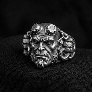 Może przedstawiać: Srebrzysty pierścień z metalu przedstawiający szczegółowo wyrzeźbioną twarz z wyraźnymi rogami, bródką i fakturowaną skórą. Boki pierścienia zdobi wzór przypominający cegły oraz motywy zwiniętych węży, wykończone ciemną, oksydowaną patyną.